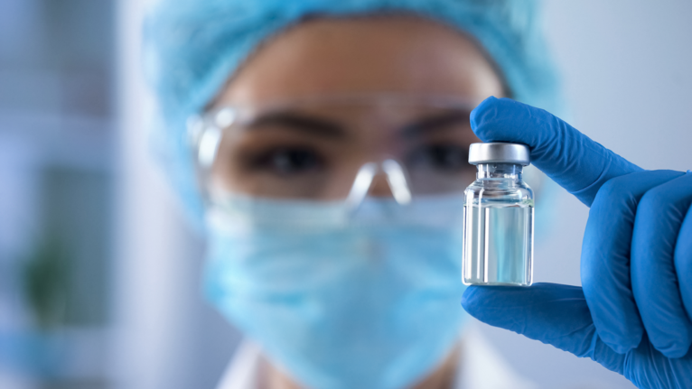 Close up of a hand wearing blue latex gloves holding a vial containing a clear liquid biopharmaceutical treatment.