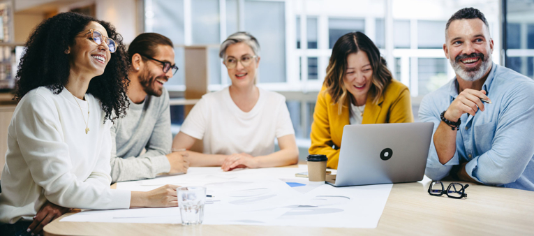 Diverse group of five professionals smiling and having a meeting around a table with documents and a laptop.
