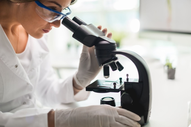 Scientist in white lab coat and gloves in a laboratory setting using a microscope to examine something.